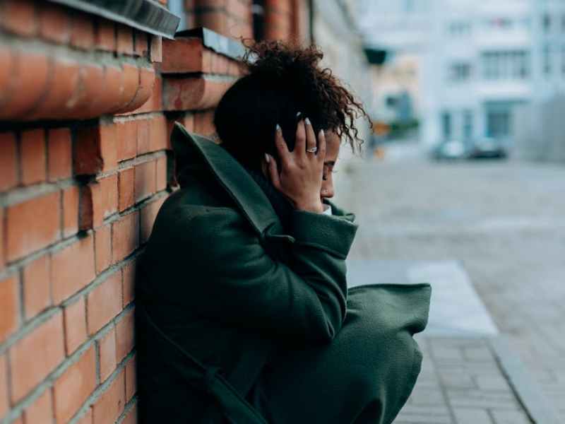 woman in green trench coat sitting and leaning against a concrete red brick wall