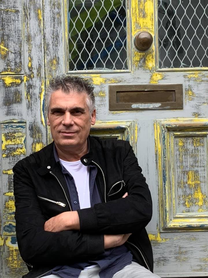 A man sitting with his arms crossed in front of a textured, colorful door, showcasing a relaxed demeanor.