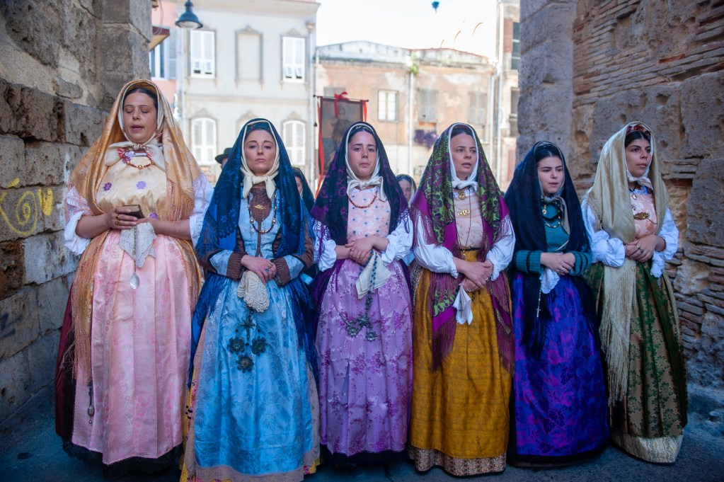 Turismo Sardegna 2026: l’isola supera i 20 milioni di presenze e guarda all’Europa dell’Est 8 Turismo Sardegna A group of women dressed in traditional colorful dresses and headscarves, standing together in a historic setting, with stone walls and vintage buildings in the background.