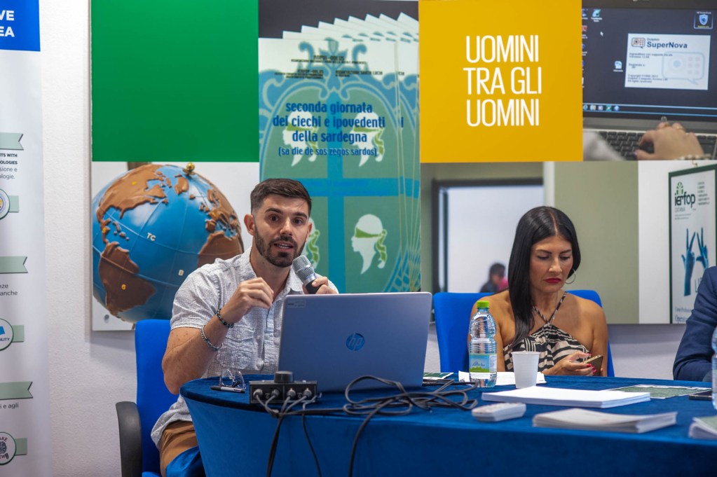 Un uomo che parla al microfono durante una tavola rotonda, mentre una donna ascolta e utilizza il telefono. Al tavolo ci sono un laptop e bottiglie d'acqua. Sullo sfondo, poster e immagini informative.