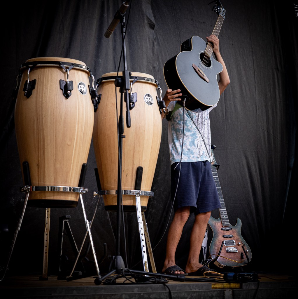 Un giovane musicista si prepara a suonare, sollevando una chitarra sopra la testa, circondato da conga e altre chitarre sul palco.