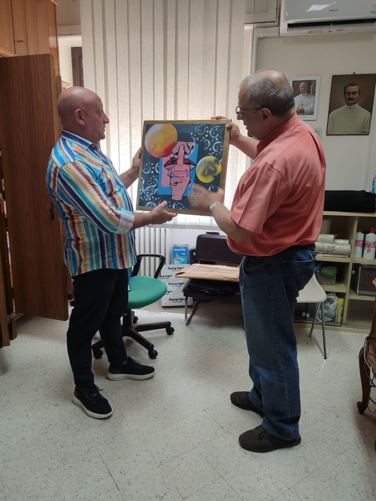 Due uomini stanno mostrando un'opera d'arte intitolata 'STOP', raffigurante una mano che silenzia, circondata da pianeti e stelle, in un ambiente interno.