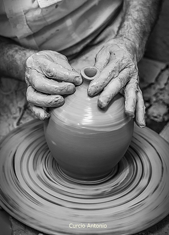 Artista che modella una forma di ceramica su un tornio, con mani visibilmente ricoperte di argilla.