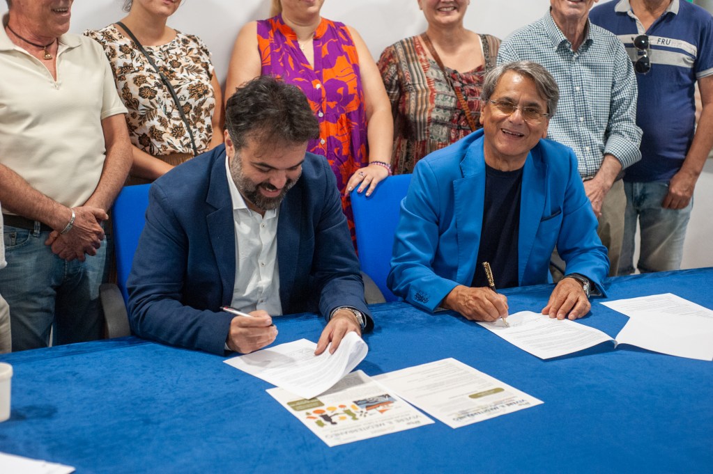 Due uomini sorridenti siedono a un tavolo coperto da una tovaglia blu, mentre firmano documenti. Dietro di loro, un gruppo di persone osserva l'evento. L'atmosfera è festosa e collaborativa.