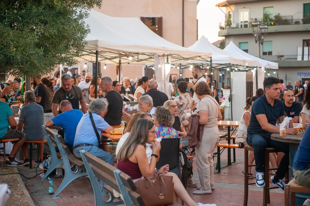 Un affollato evento in piazza durante la Sagra de Sa Panada ad Assemini, con visitatori seduti attorno a tavoli e stand che offrono specialità locali.