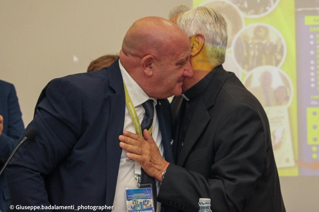 Due uomini si abbracciano con affetto durante un evento, uno indossa un abito scuro e l'altro un abito priest, lo sfondo mostra immagini di un convegno.