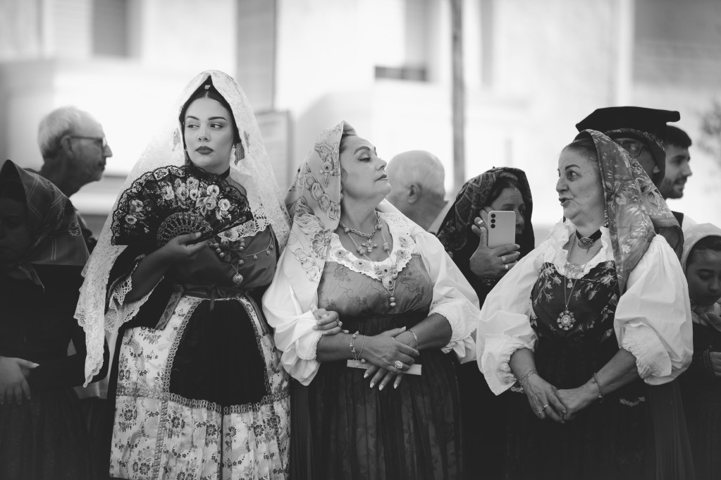 Gruppo di donne in abito tradizionale sardo sfila per le vie di Assemini durante la Sagra de Sa Panada 2025, accompagnate dalla musica popolare. Immagine in bianco e nero dal forte impatto emotivo