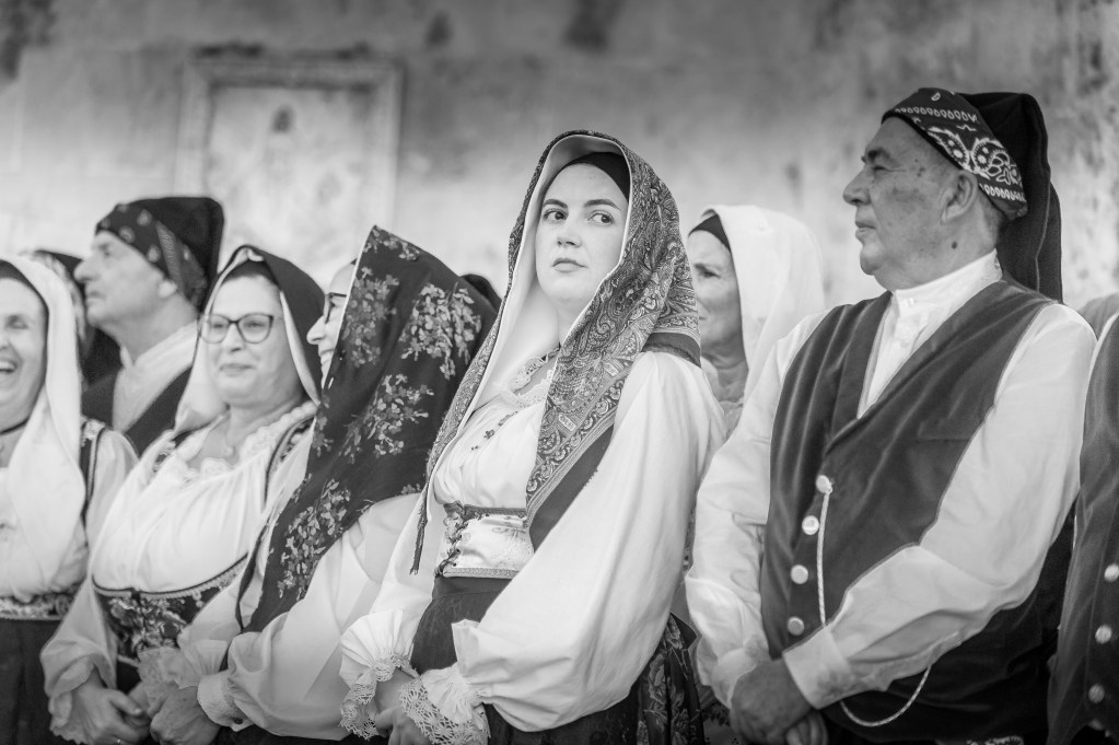 Gruppo di donne in abito tradizionale sardo sfila per le vie di Assemini durante la Sagra de Sa Panada 2025, accompagnate dalla musica popolare. Immagine in bianco e nero dal forte impatto emotivo