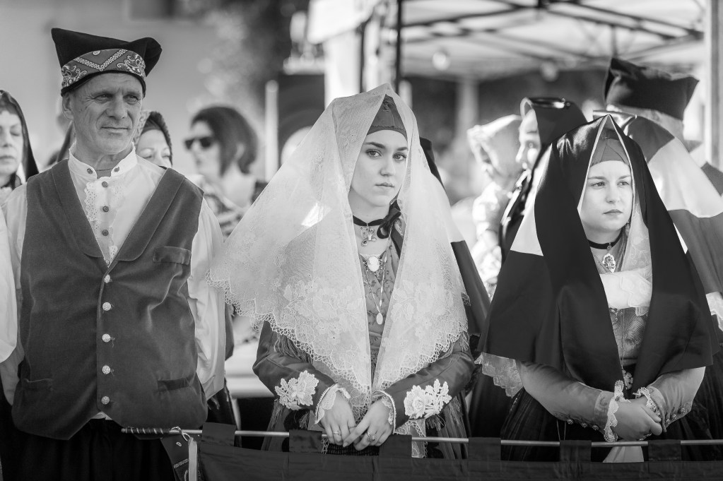 Due donne in abiti tradizionali sardi con corone di pizzo e un uomo in costume tradizionale, tutti in una manifestazione folkloristica, in bianco e nero.