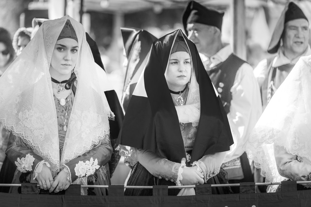 Gruppo di donne in abito tradizionale sardo sfila per le vie di Assemini durante la Sagra de Sa Panada 2025, accompagnate dalla musica popolare. Immagine in bianco e nero dal forte impatto emotivo