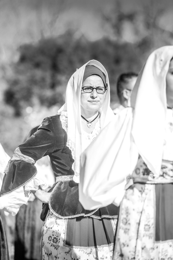 Una donna in costume tradizionale sardo durante la Sagra de Sa Panada, con un'espressione seria e attenta, mentre partecipa a una sfilata folkloristica.