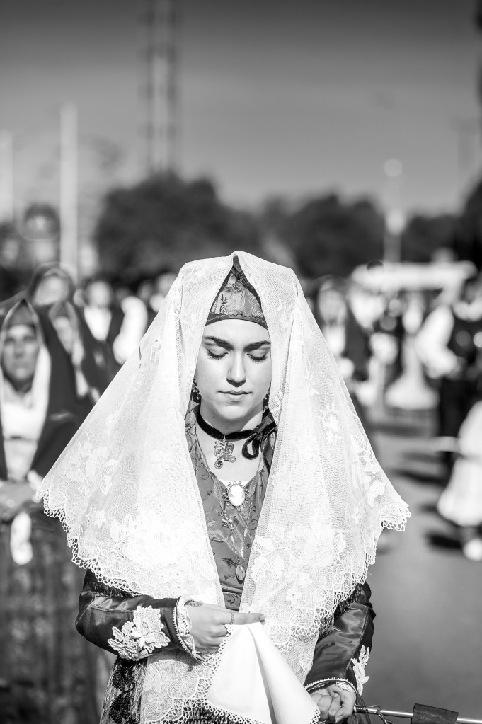 Giovane donna in costume tradizionale sardo durante la sfilata folkloristica della Sagra de Sa Panada 2025 ad Assemini. Ritratto in bianco e nero che cattura fierezza ed eleganza.