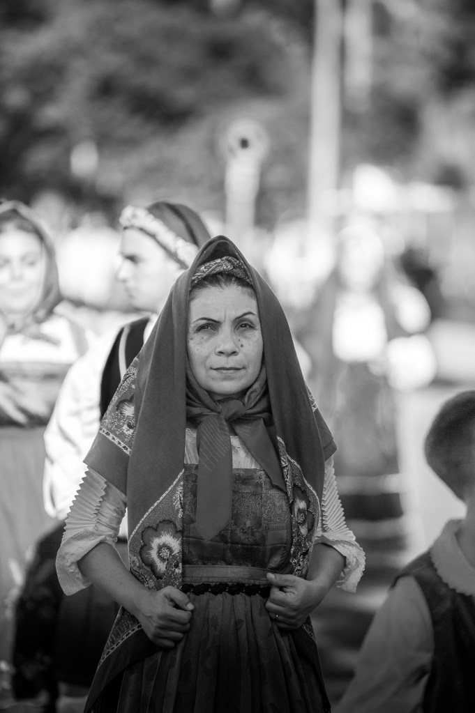 Donna anziana in abito tradizionale sardo durante una sfilata folkloristica, con un'espressione di fierezza e solemnità.