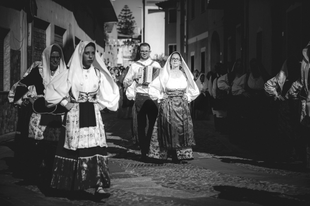 Sfilata di persone in costumi tradizionali sardi durante la Sagra de Sa Panada, con un musicista che suona l'organetto in un vicolo, in bianco e nero.