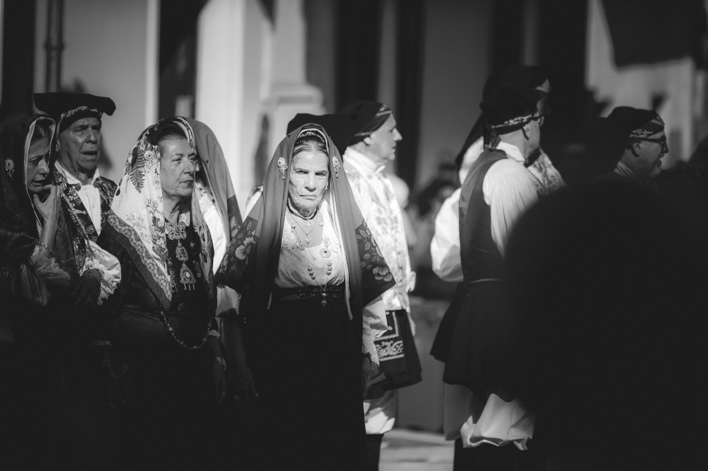 Un gruppo di persone in costumi tradizionali sardi durante una sfilata alla Sagra de Sa Panada, catturati in un'immagine in bianco e nero.
