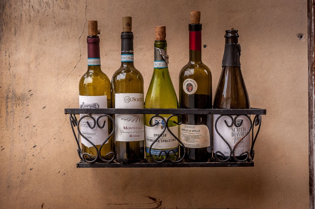 A metal wine rack displaying five bottles of wine against a textured wall in Spello, Perugia.