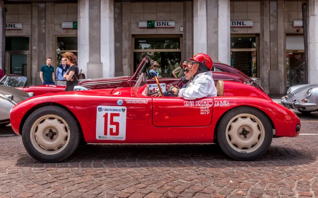 A classic red sports car parked on a cobblestone street, featuring a driver wearing a white outfit and a red helmet, holding a map. The car displays the number 15 and has the event name 'Gran Premio di Bider' on its side.