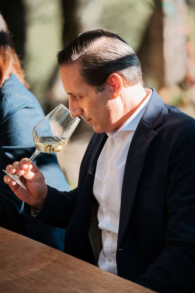 A man in a blazer studies a glass of Vermentino wine, holding it close as he inhales its aroma. The setting has a natural outdoor backdrop.