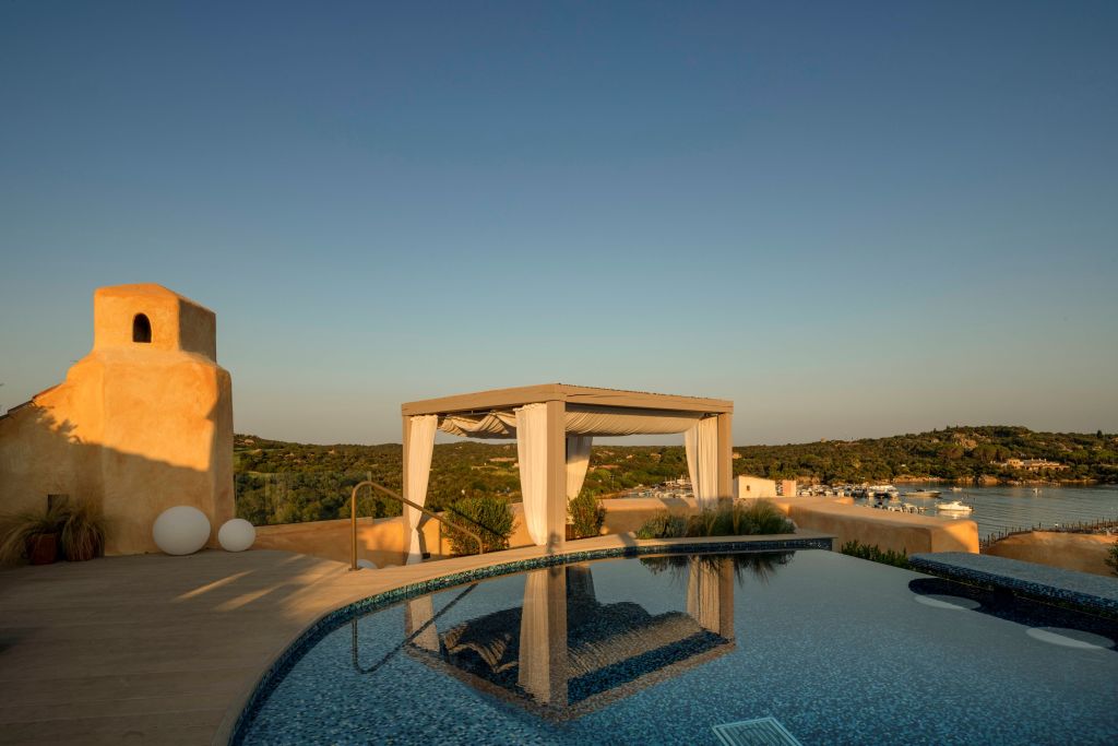 View of a modern rooftop pool area with a pergola overlooking a scenic coastline at sunset.