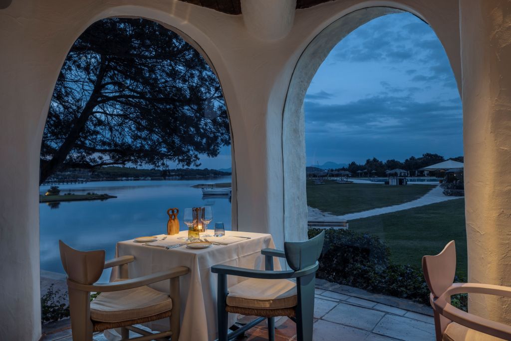 A serene dining setting viewed through arched windows, with a table set for two, overlooking a calm body of water at dusk, a tree visible in the foreground.
