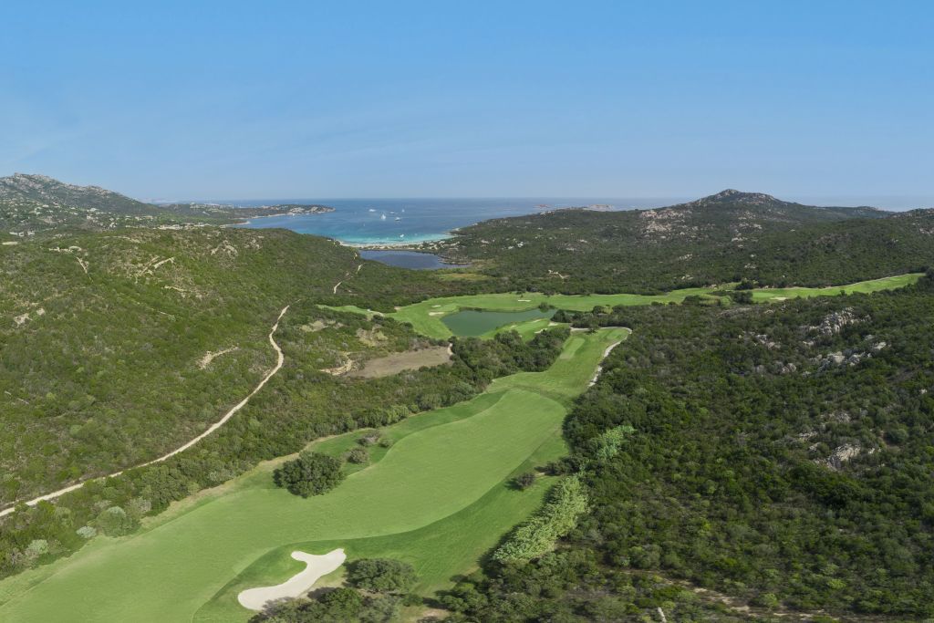 Aerial view of a lush green golf course surrounded by hills, overlooking a blue coastal sea.